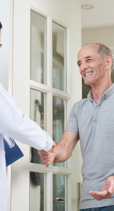Female doctor with clipboard shaking hand of senior man during home visit while entering house